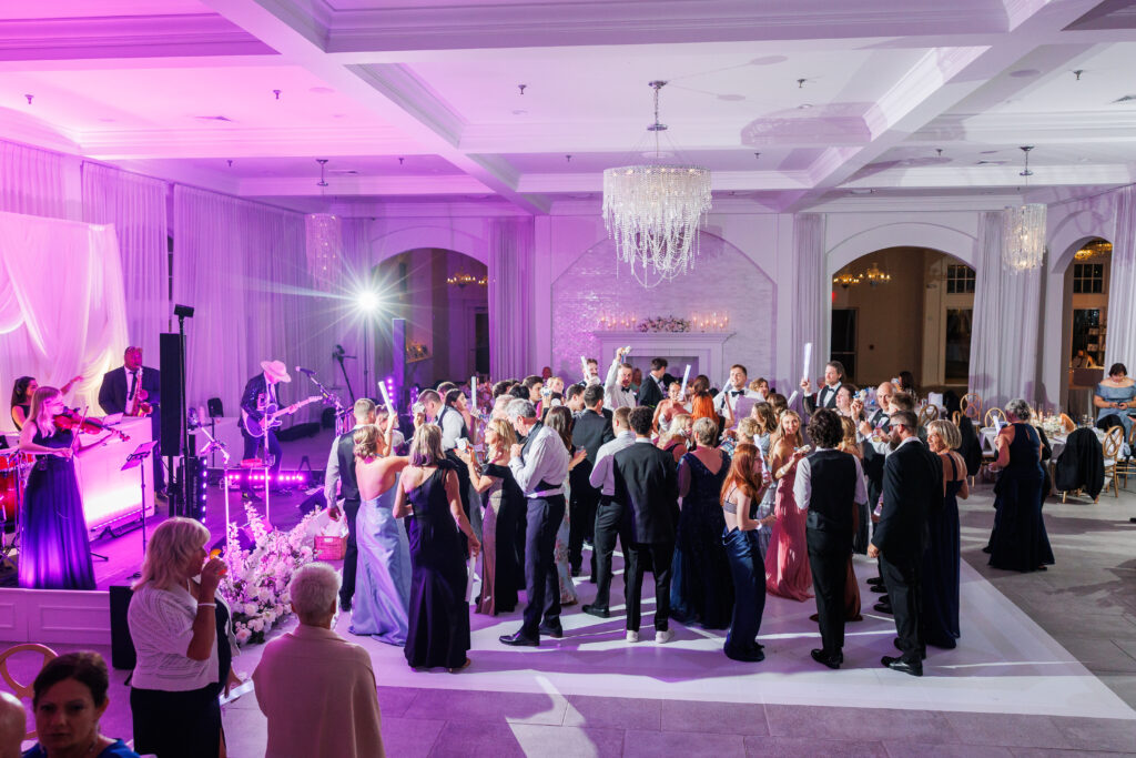 Wedding reception with a live band performing on the left while guests dance on a pink-lit ballroom floor.