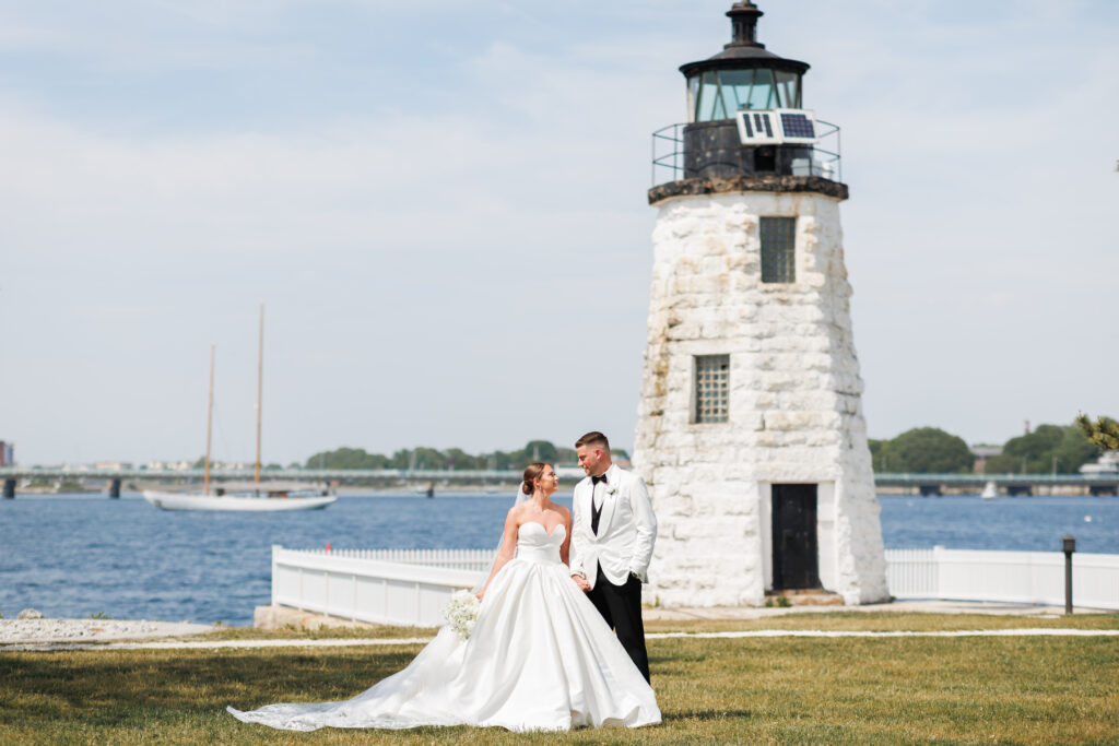 Bride and groom in wedding attire stand hand in hand near a white stone lighthouse by the water, with a sailboat in the background.